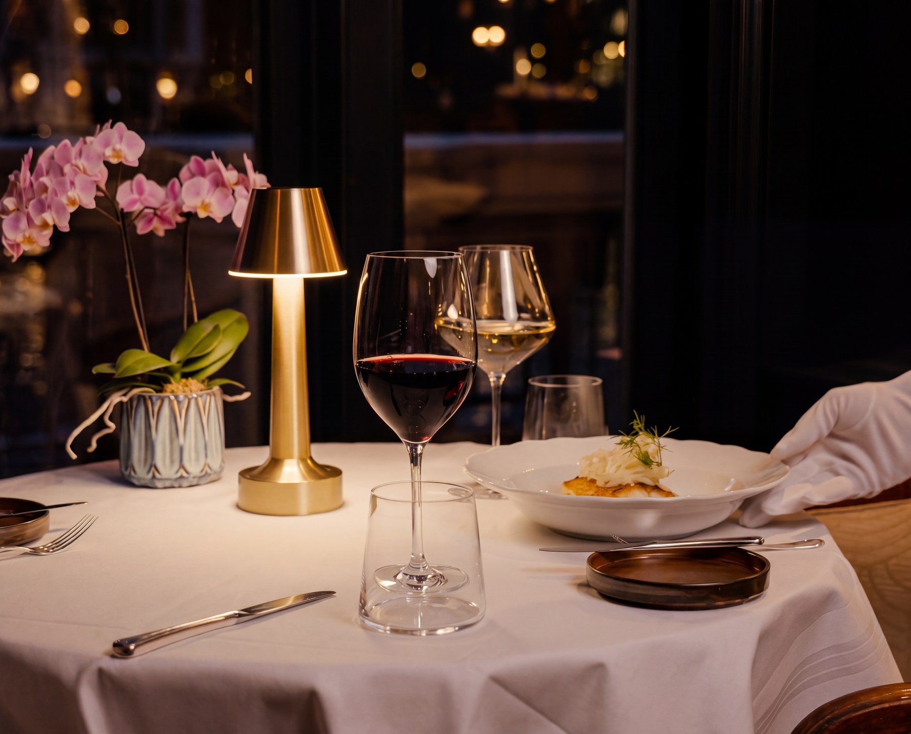 A dressed table lit by a brass Clarté lamp, a waiter placing a plate.