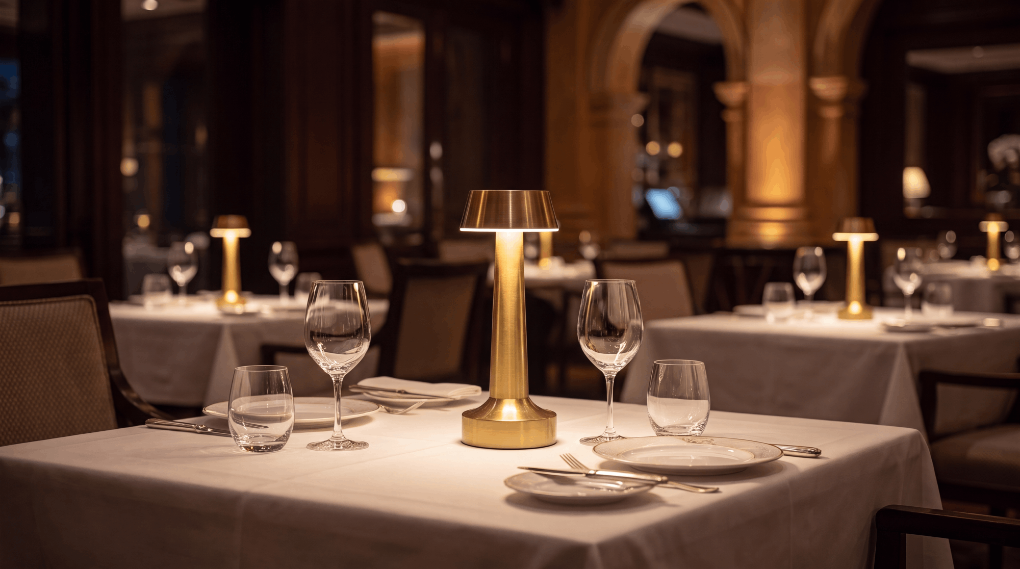 A restaurant table set with a cordless brass Clarté lamp, wine glasses and white linen in a warm columned dining room